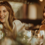 Duas mulheres conversam e riem durante um evento corporativo, num ambiente elegante e acolhedor com luz natural